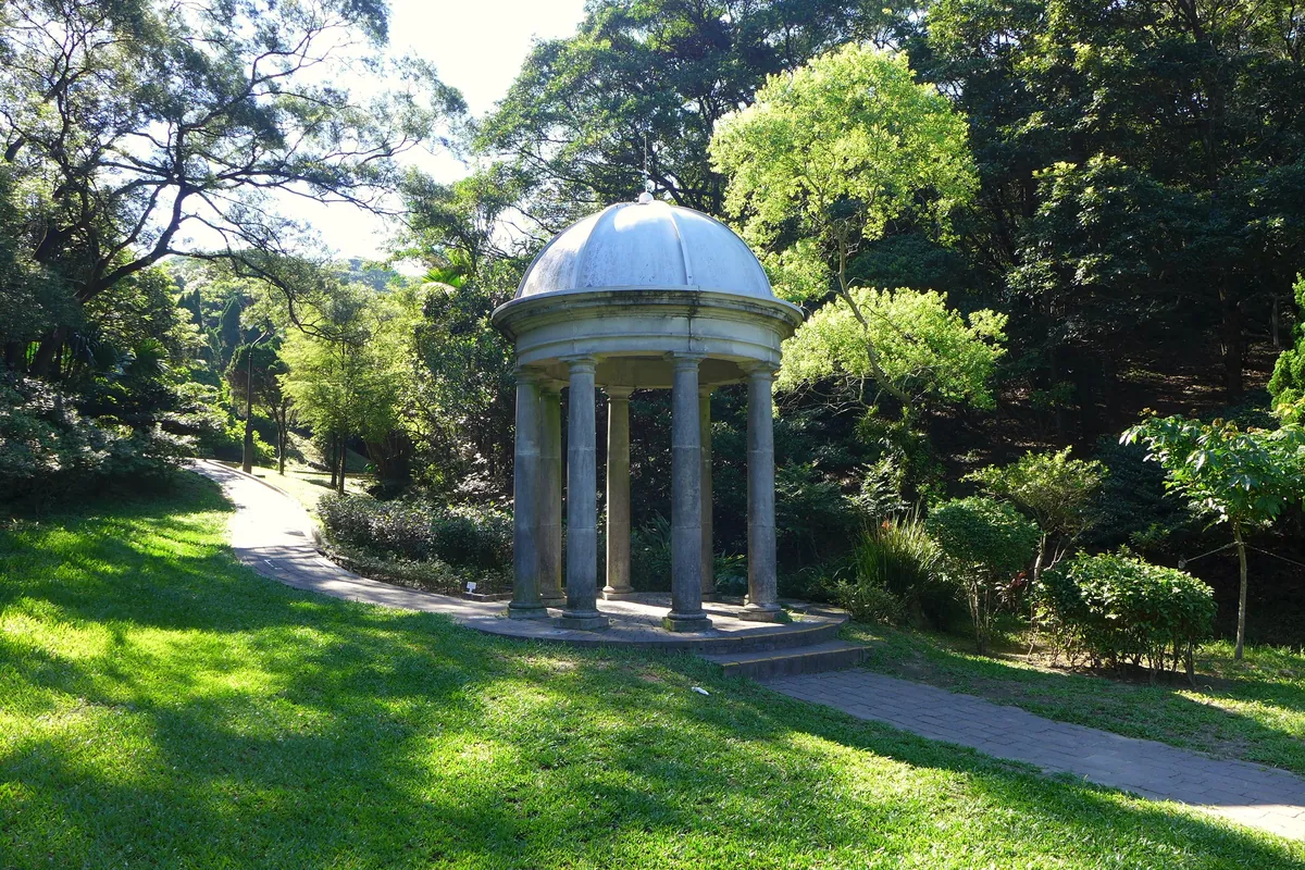 Ein steinerner Pavillon mit Kuppeldach auf einer Rasenfläche im Victoria Peak Garden, umgeben von Bäumen