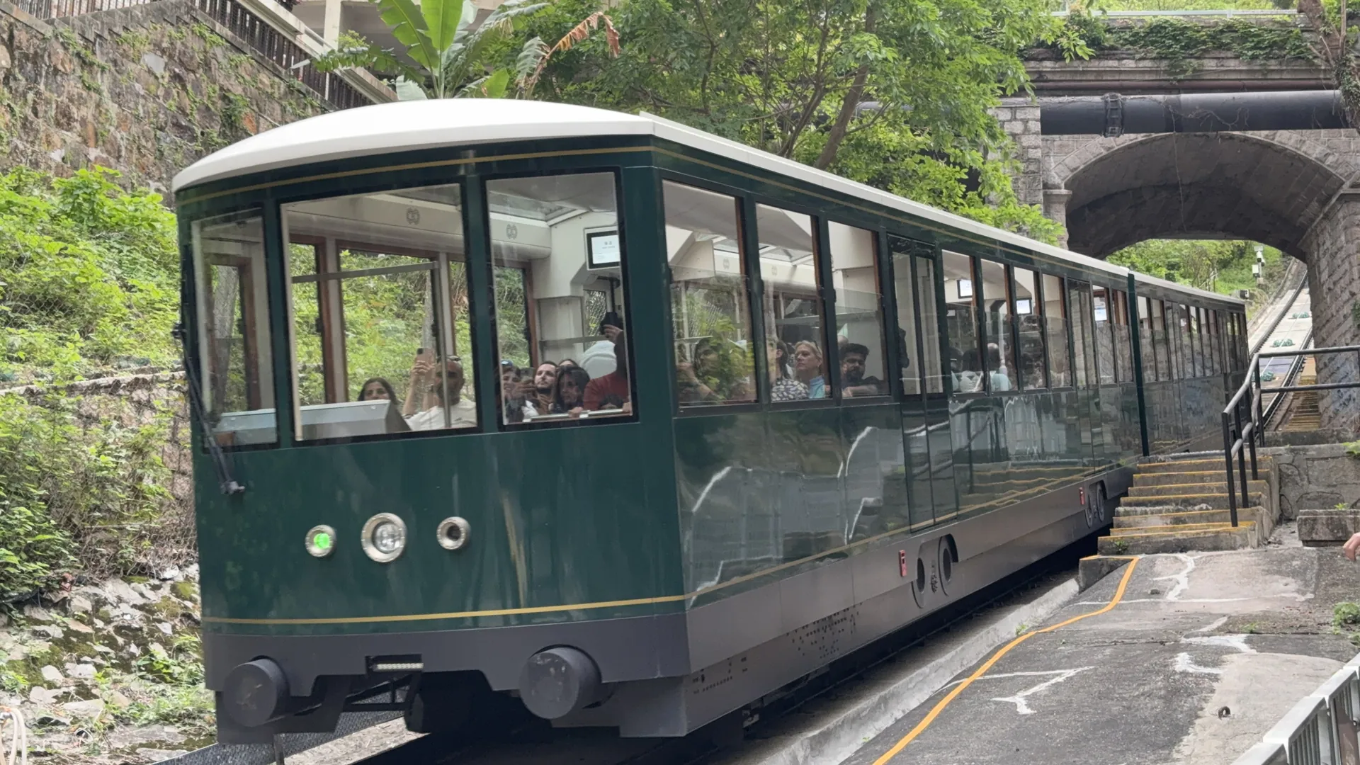 A green Peak Tram car coming out of a stone arch on its climb towards Victoria Peak, with passengers on board